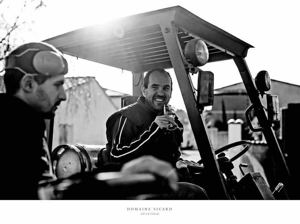 Deux vignerons du Domaine Sicard à Aigues-Vives, en pleine discussion près d’un tracteur, l’un d’eux tenant un verre de vin, illustrant l’ambiance conviviale et artisanale d’un domaine viticole familial du Minervois.