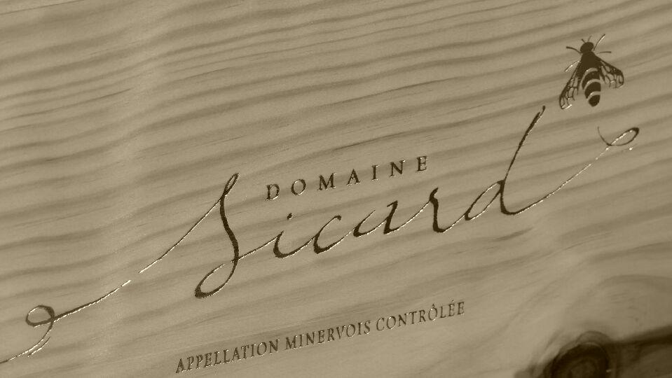 Logo gravé du Domaine Sicard, producteur de vin Minervois AOC, avec une abeille stylisée et mention “Appellation Minervois Contrôlée” sur une caisse en bois.